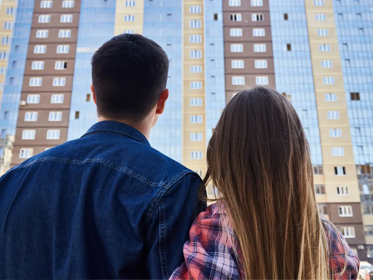 A man and a woman, seen from behind, stand close together and look up at a tall, modern apartment building with many windows.