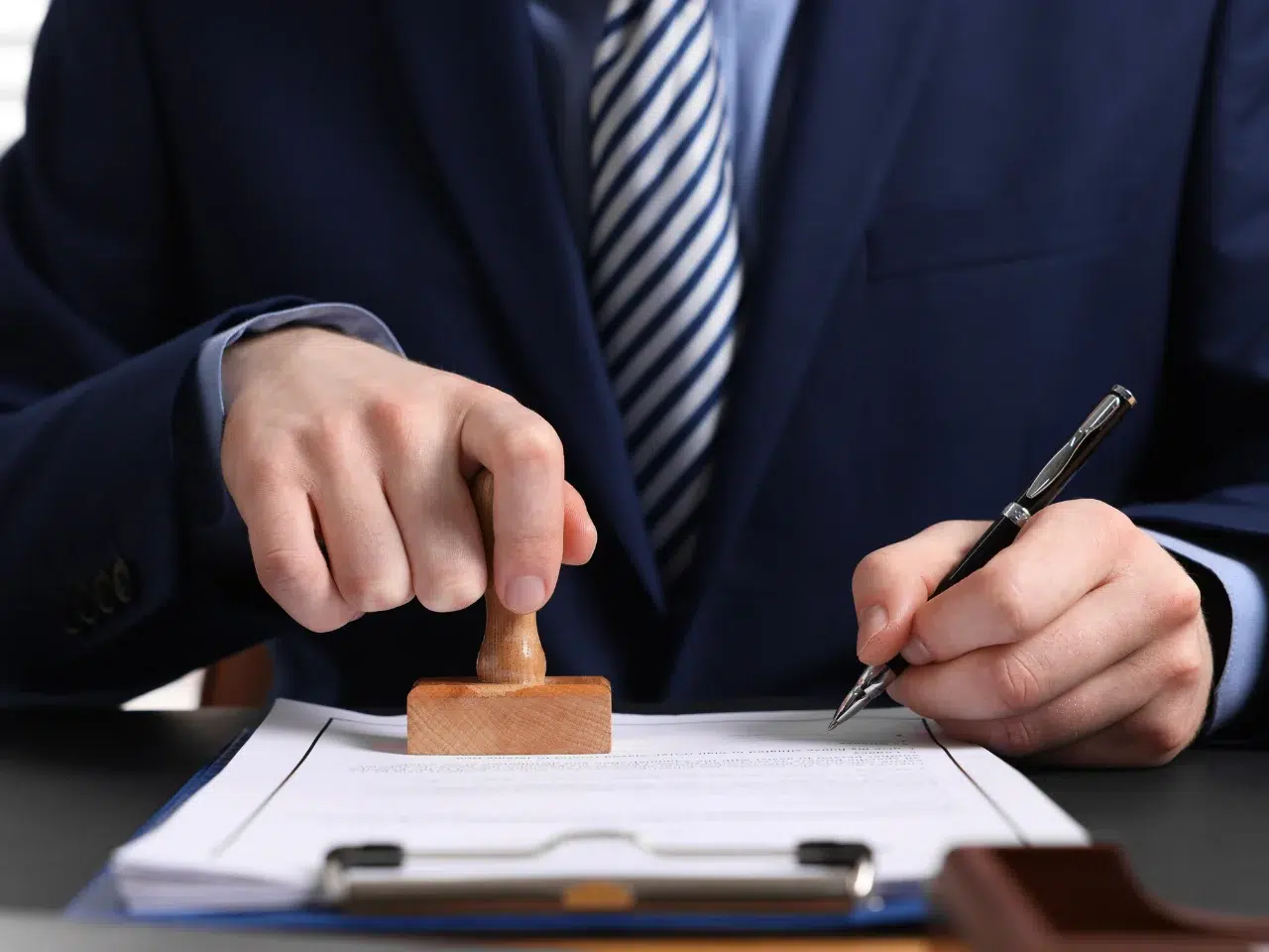 A person in a suit stamps a document while holding a pen, sitting at a desk with paperwork and a clipboard.