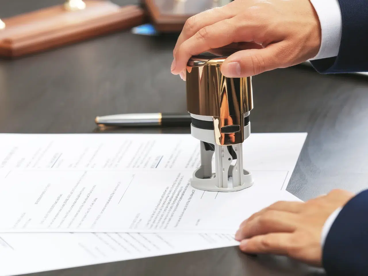 A person presses a round stamp onto a document on a desk, with a pen and another document visible in the background. The scene suggests official approval or authorization of paperwork.