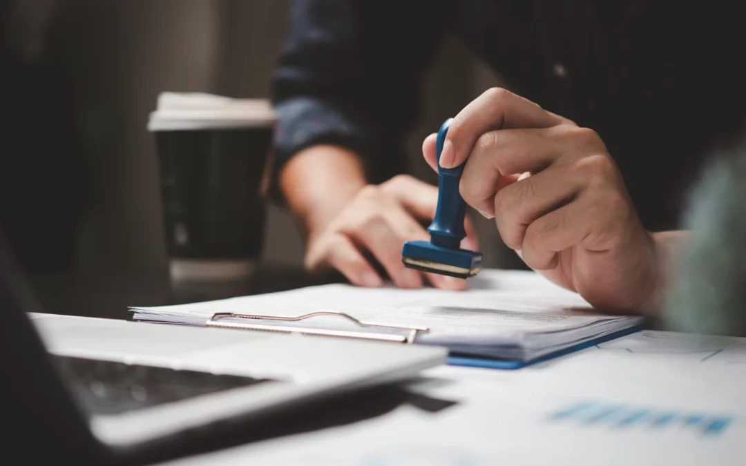 A person presses a blue stamp onto a document on a clipboard, with a laptop and a disposable coffee cup nearby, suggesting an office or business setting.