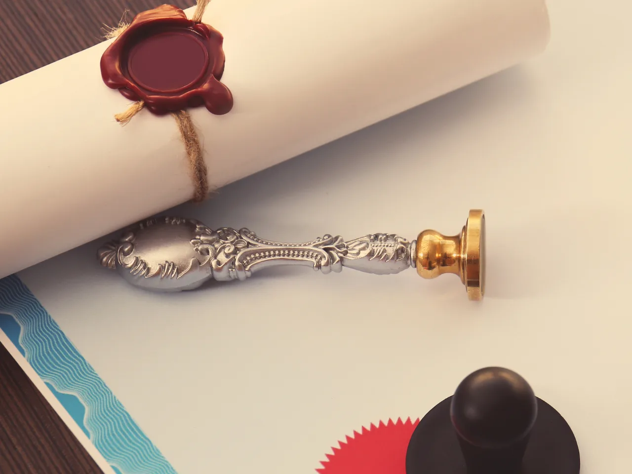 A decorative metal wax seal stamp, a rolled parchment tied with twine and sealed with red wax, and certificate papers with embossed seals are arranged on a light surface.