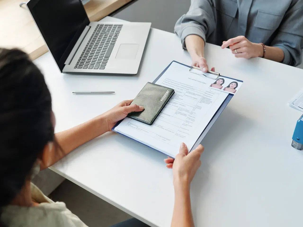 Two people sit at a desk. One hands a clipboard with a form and passport to the other. A laptop, pen, and documents are on the table, suggesting a formal meeting or identity verification process.
