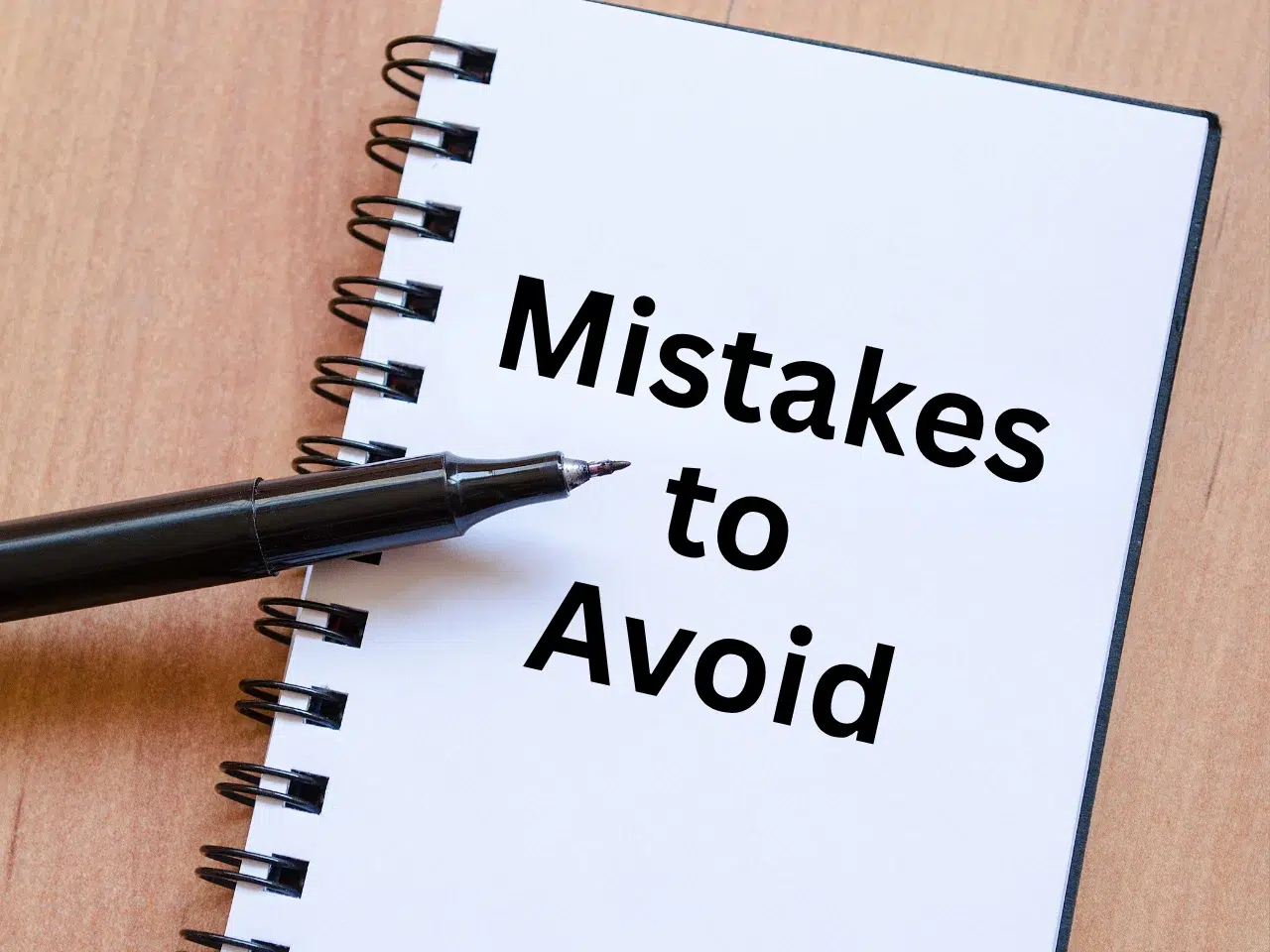 A black pen rests on an open spiral notebook with the words Mistakes to Avoid written in bold black letters on a white page. The notebook is placed on a light wooden surface.