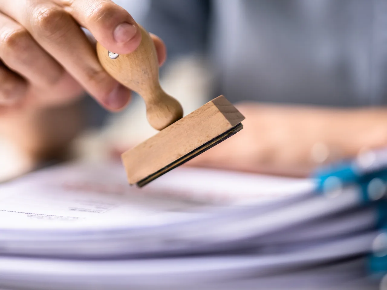 A close-up of a hand pressing a wooden stamp onto a stack of documents, with paperwork and a blue clip visible in the foreground.