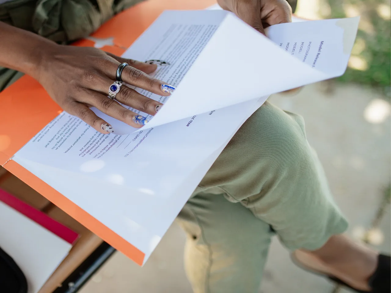 A person with patterned nails and rings flips through printed documents in an orange folder while sitting cross-legged outdoors.