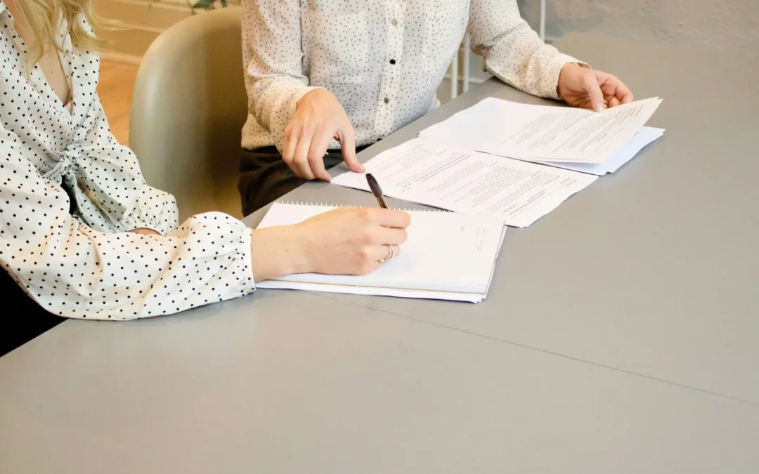 Two people sitting at a table reviewing and discussing documents. One person is holding a pen over the papers, while the other points at the documents. Both are wearing light-colored, patterned shirts.