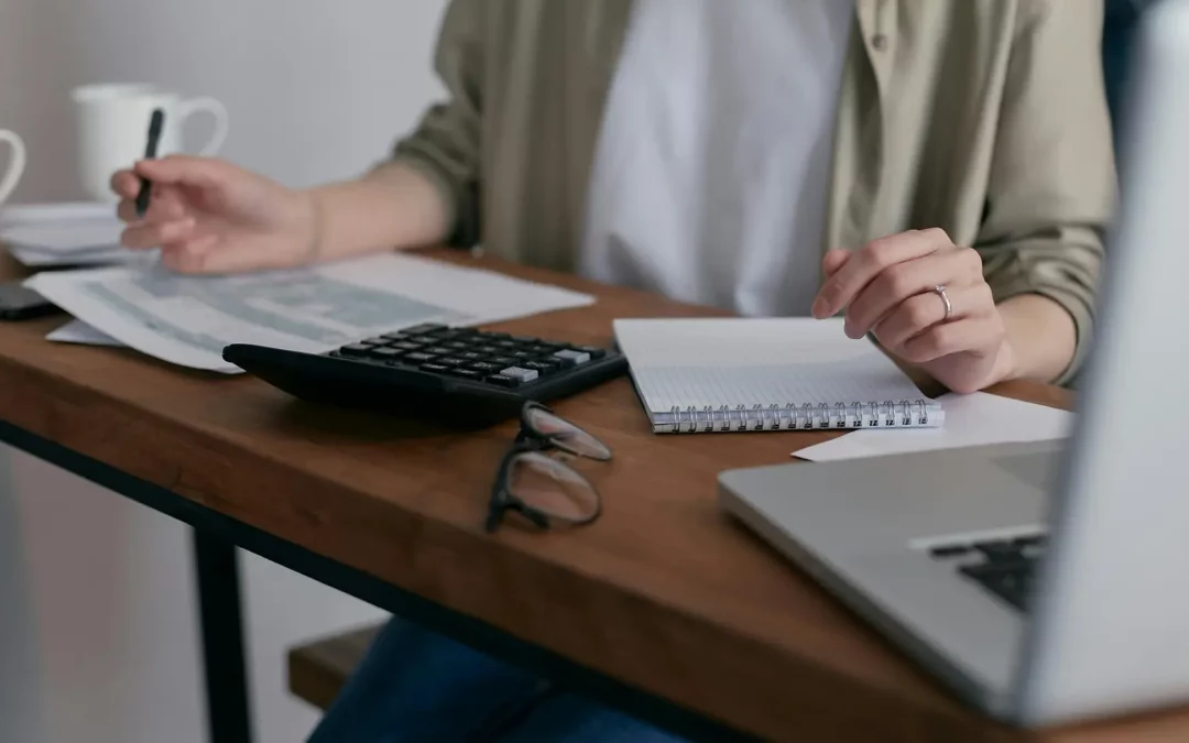 A person sits at a desk with a laptop, notebook, calculator, glasses, and papers, appearing to work or study. Two coffee mugs are on the table. Only the persons torso and hands are visible.