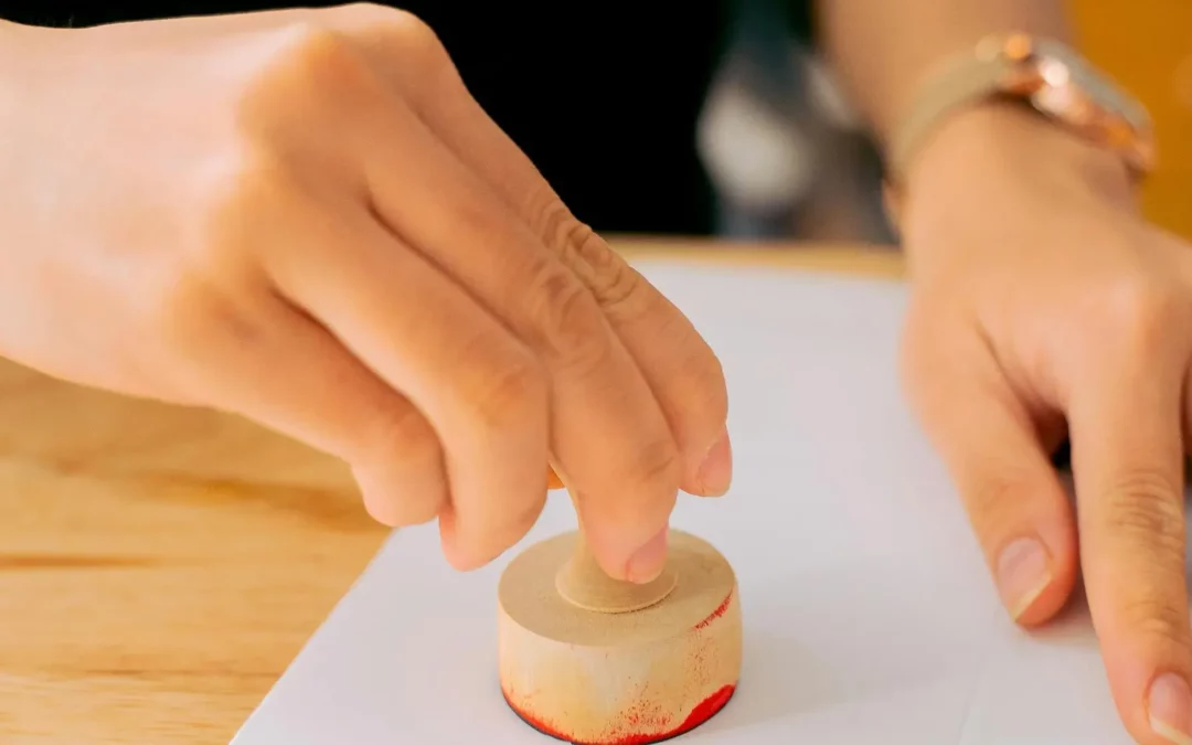 A close-up of a person’s hand pressing a round wooden stamp with red ink onto white paper, with another hand holding the paper steady on a wooden table.