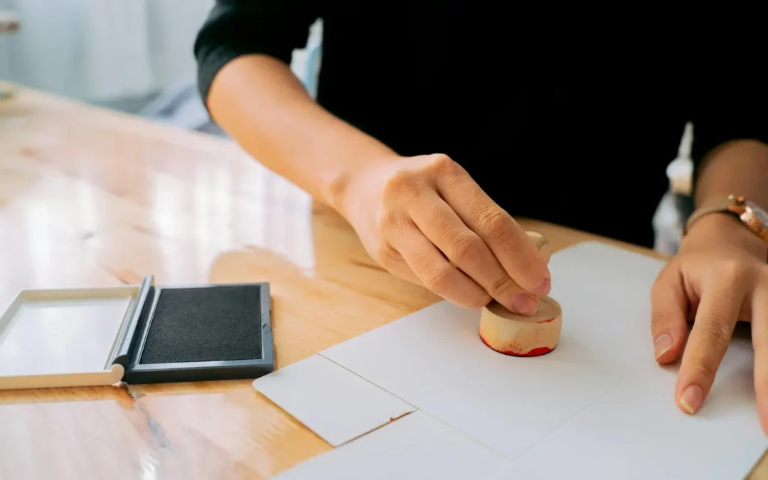 A person presses a round wooden stamp onto a piece of paper, with a black ink pad nearby on a wooden table.