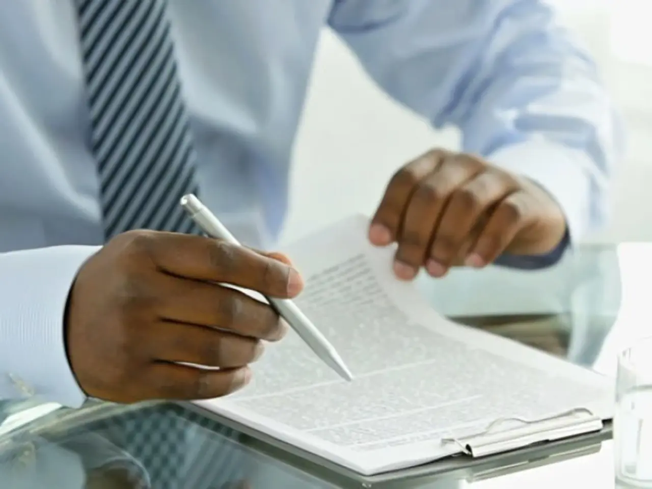 A person wearing a striped tie and light blue shirt is holding a pen and reviewing documents or papers on a glass desk.