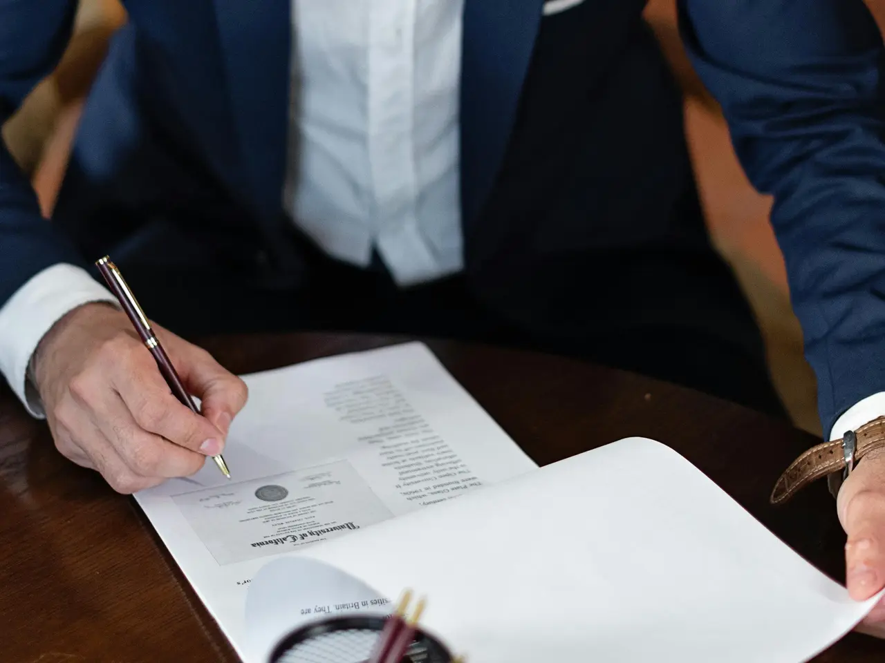A person in a suit sits at a desk, signing a document with a pen. The paper appears to be an official form or contract, and a pen holder is visible in the foreground.