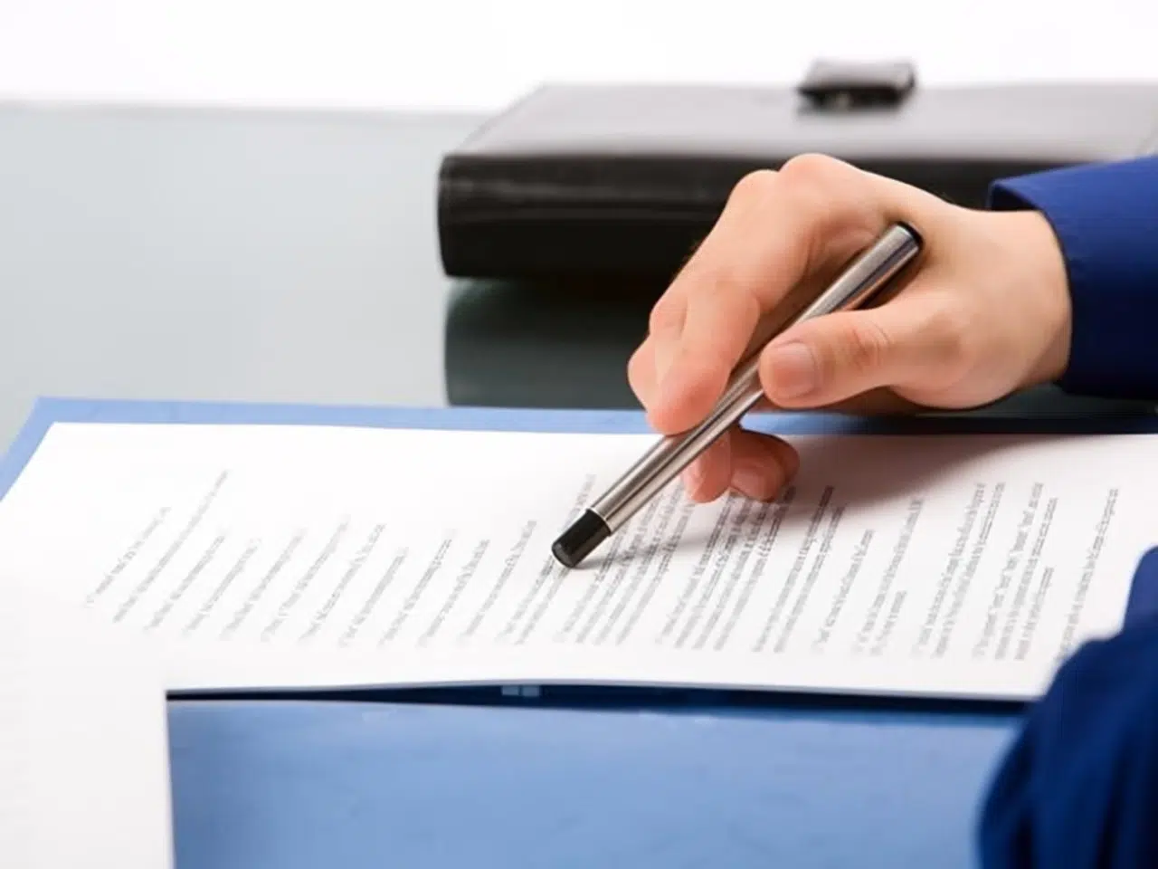 A person in a blue sleeve holds a pen and points to text on a printed document placed on a desk, with a closed black folder in the background.