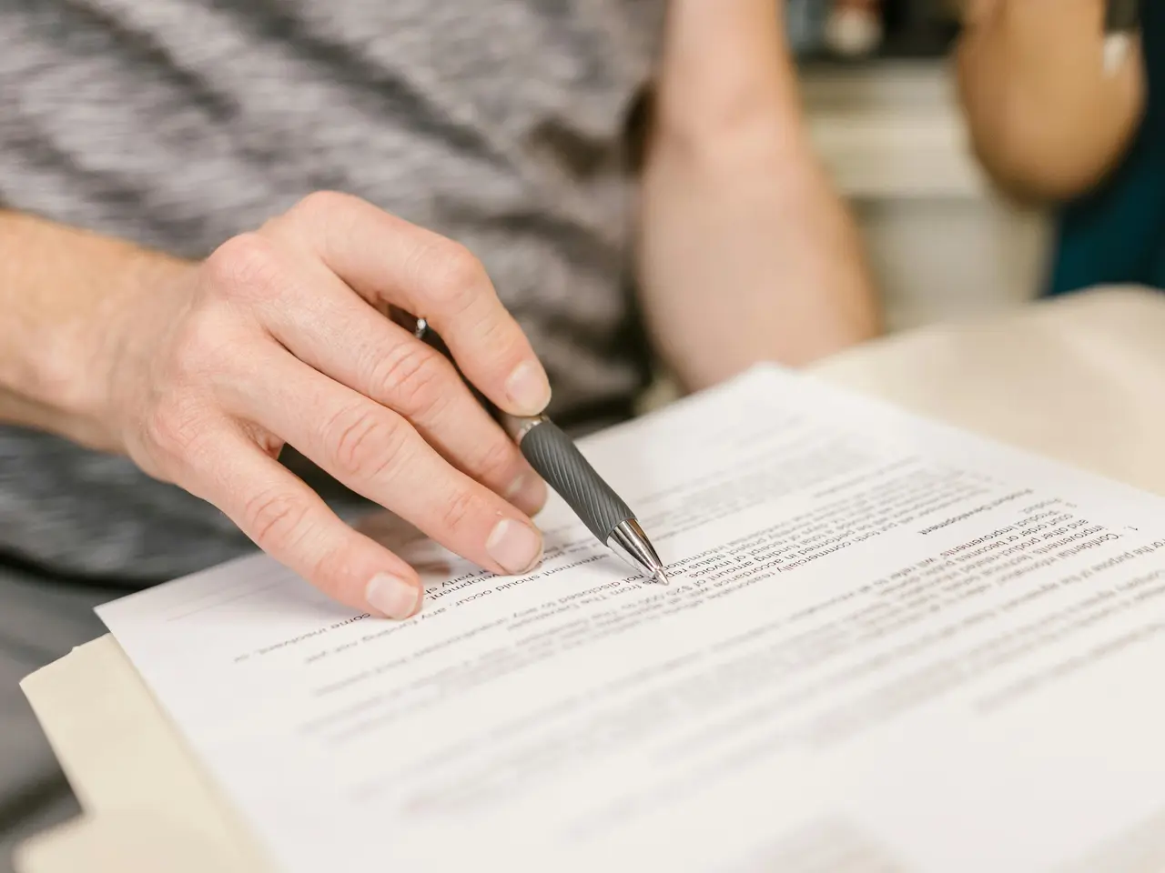 A person holds a black pen and points to text on a printed document, appearing to review or sign paperwork. The background is blurred, focusing attention on the hand and document.