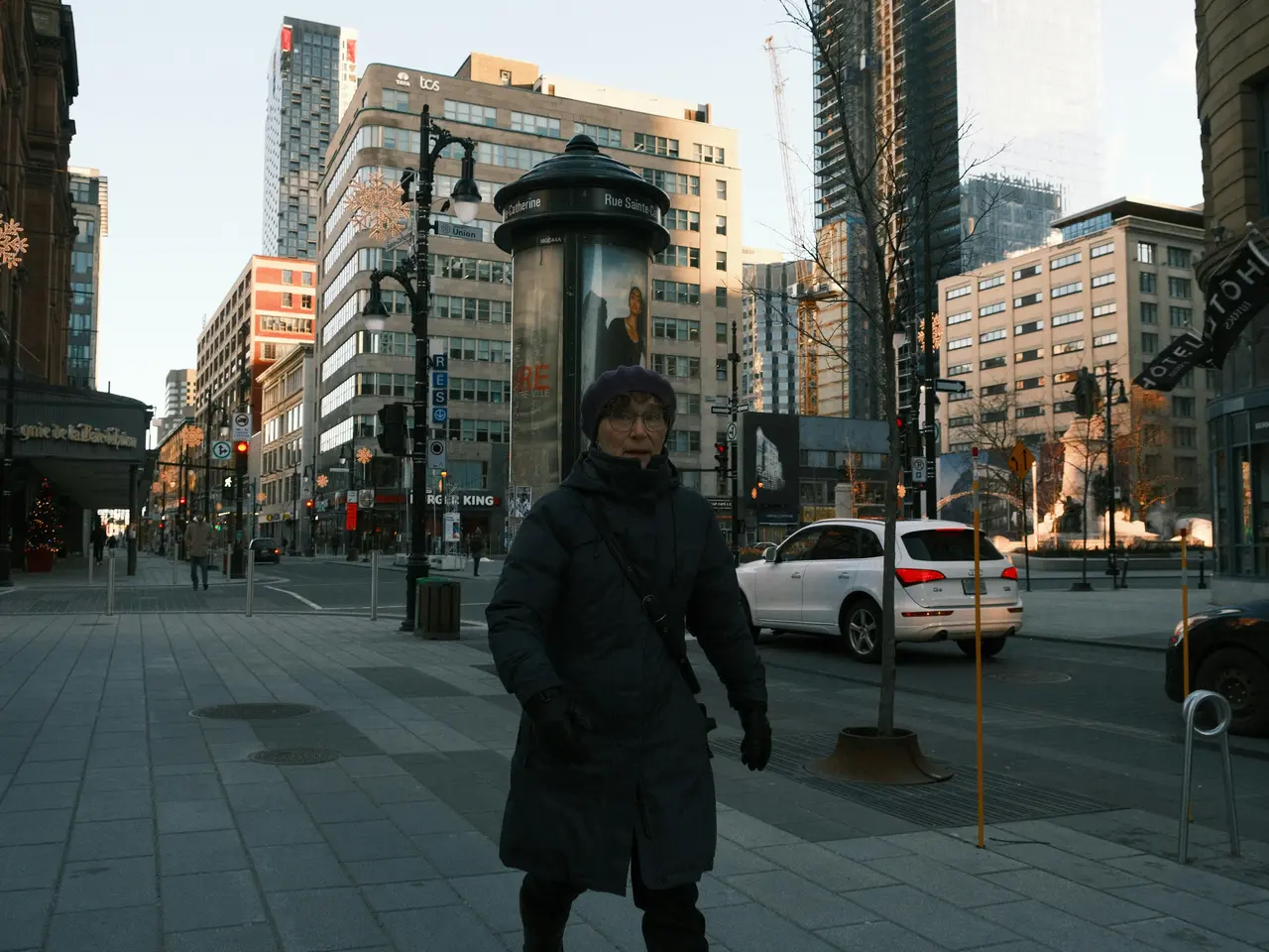 A person in a dark winter coat and hat walks on a city sidewalk lined with tall buildings, streetlights, and a few cars parked along the road on a cold, sunny day.