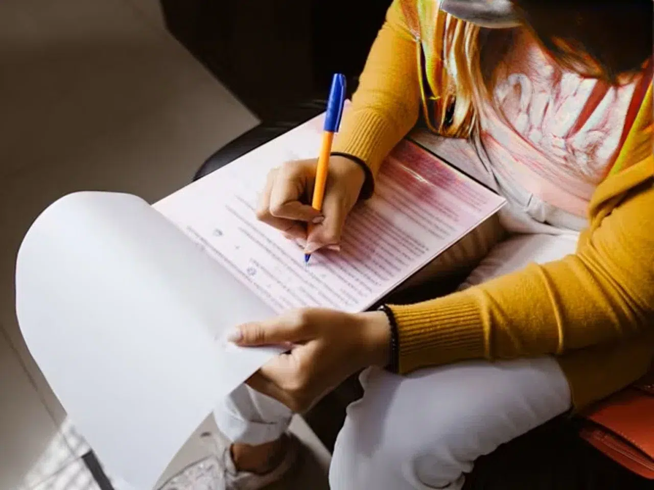 A person wearing a yellow sweater and white pants is filling out a form with a blue pen while holding the paper on their lap. Their face is not visible, and they appear to be sitting indoors.