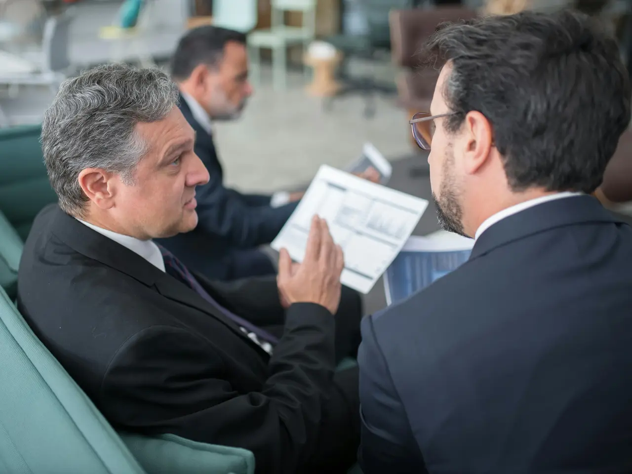 Two men in suits are seated and engaged in a serious discussion, one holding a document with charts. Another man in the background is also looking at papers, suggesting a business meeting or conference setting.