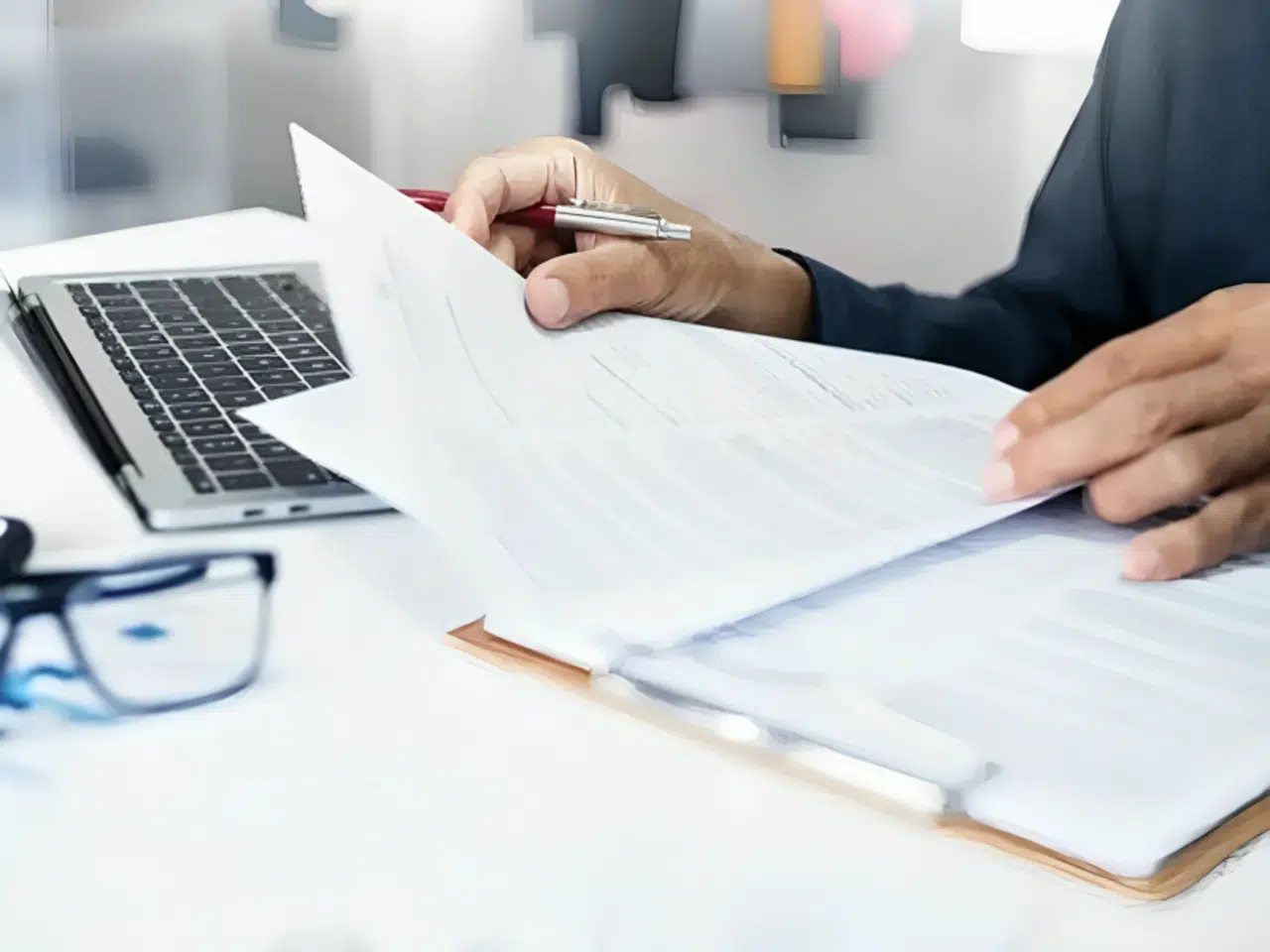 A person holding a pen reviews documents on a clipboard beside an open laptop, with a pair of eyeglasses resting on the desk in the foreground.