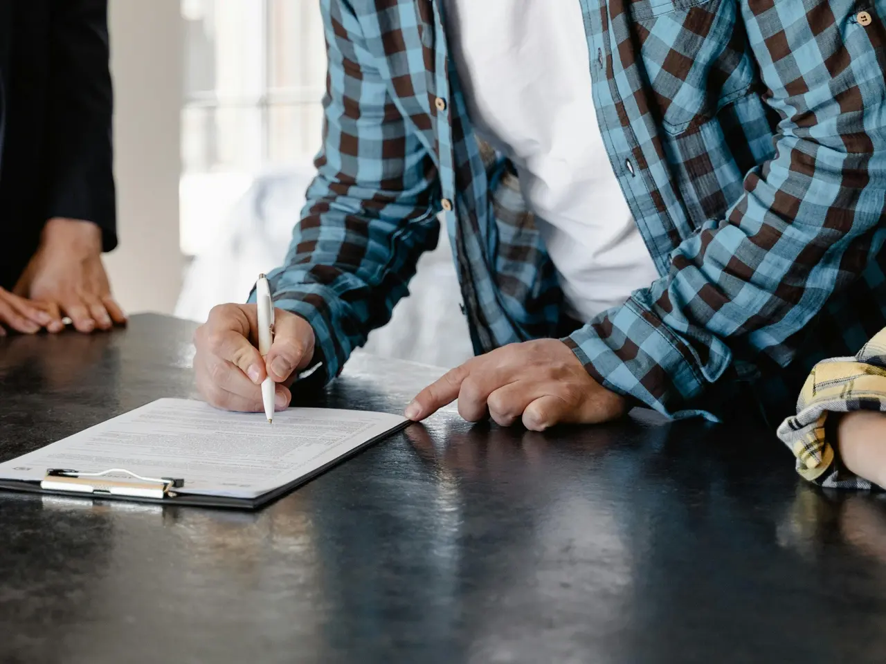 A person wearing a blue plaid shirt signs a document on a clipboard while standing at a dark table. Another individual stands nearby with only their hands visible.
