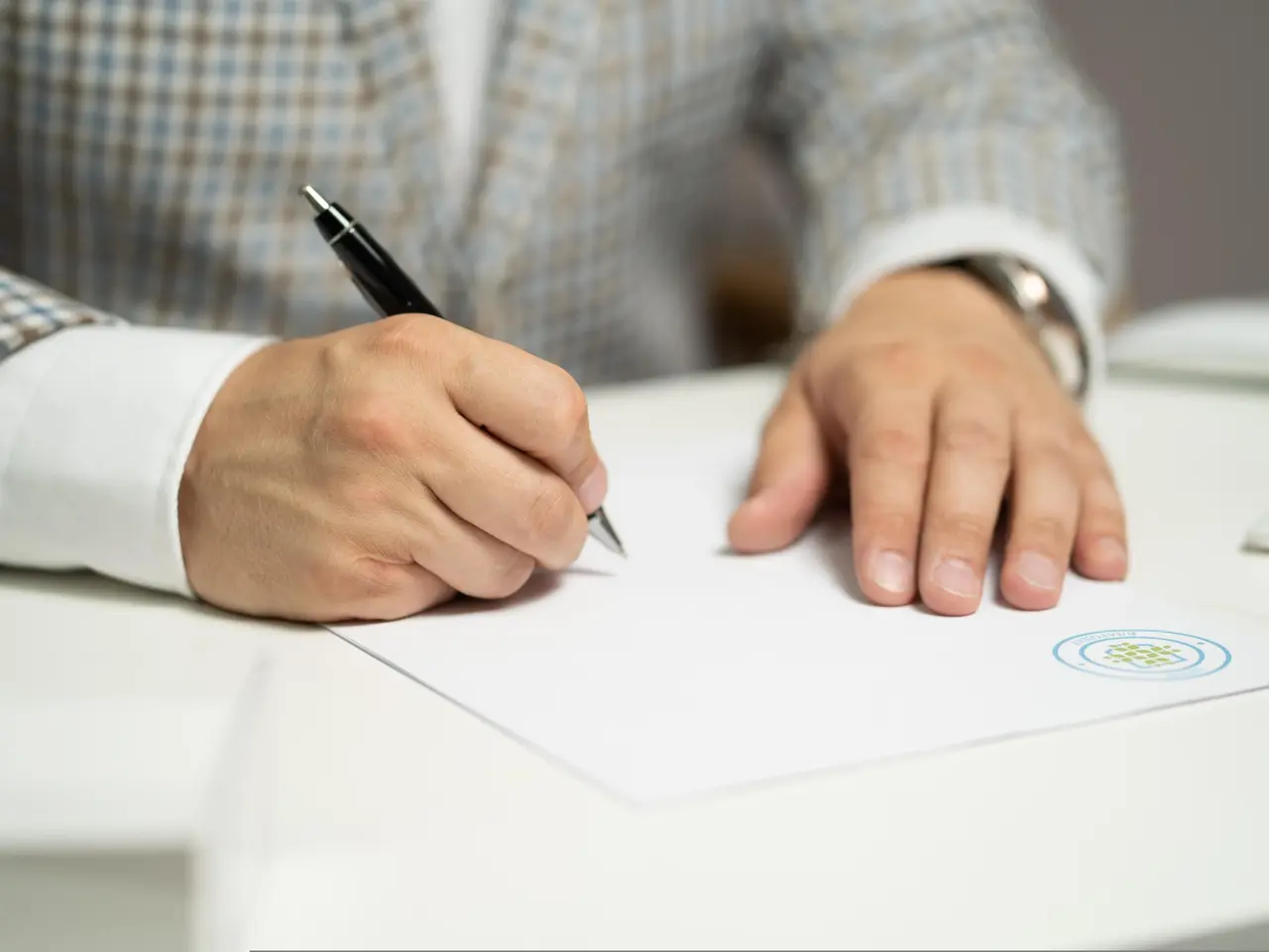 A person in a plaid jacket is writing on a blank sheet of paper with a black pen at a white desk. Only their hands and part of the torso are visible.