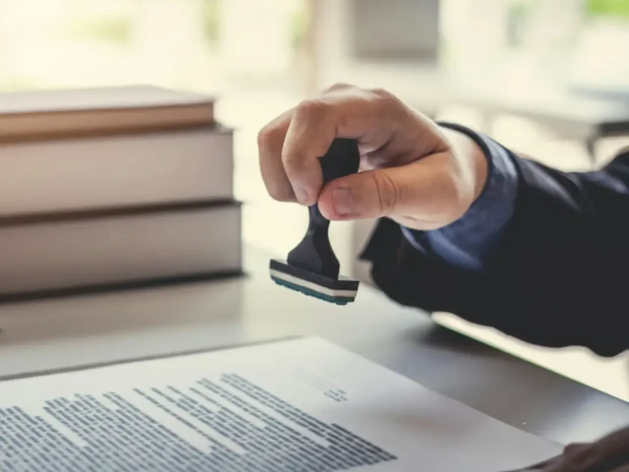 A person’s hand presses a rubber stamp onto a document on a desk, with a stack of books in the background. The scene suggests approval or official authorization of paperwork.