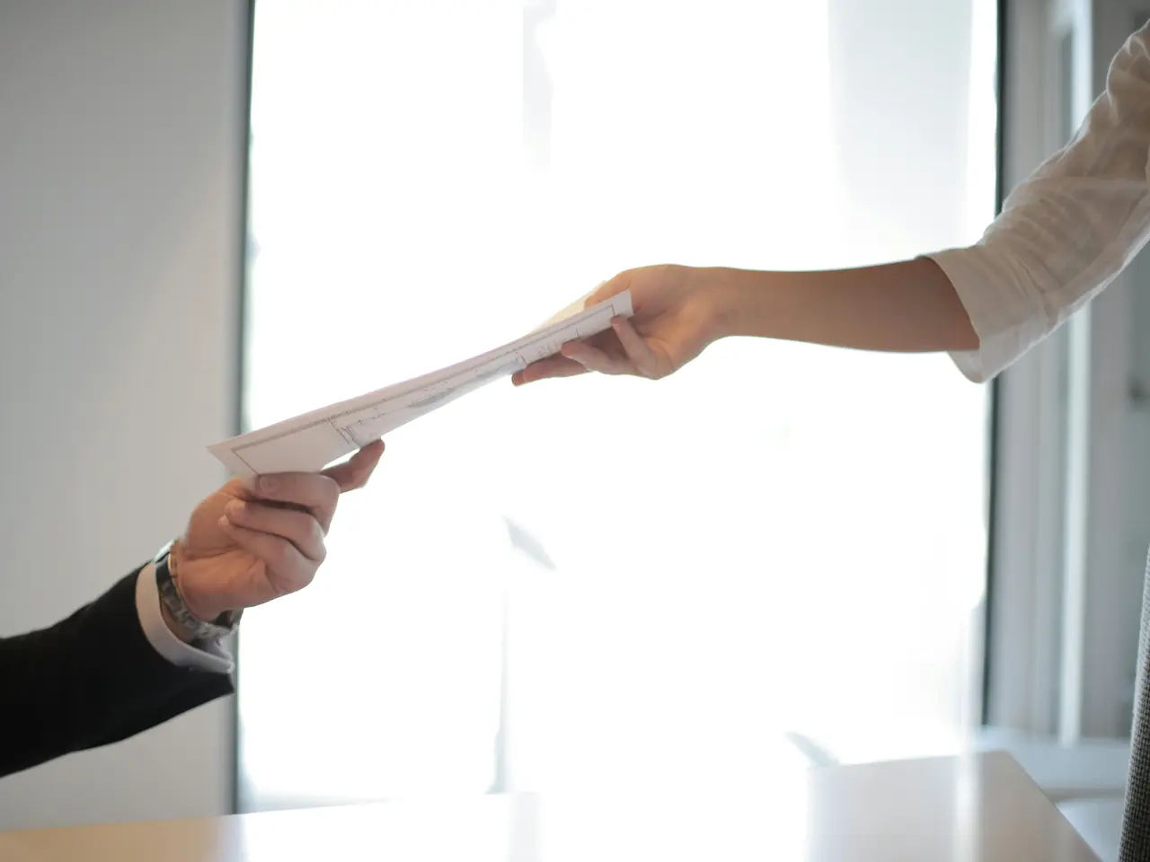 Two people exchanging a stack of papers; one person hands the documents to another in a bright, well-lit setting with a white background. Only their arms and hands are visible.
