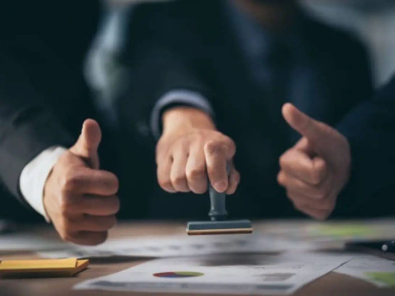 Three people in business attire sit at a table; the person in the center stamps a document while the other two gesture thumbs up, suggesting approval or agreement. Papers and charts are visible on the table.