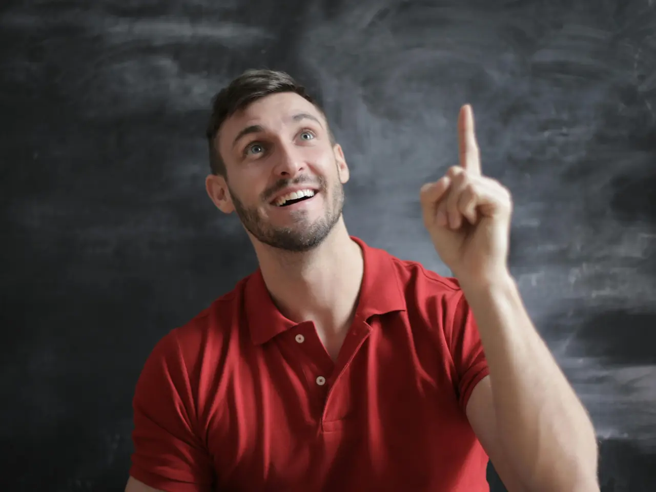 A smiling man in a red polo shirt points upwards with his index finger, standing in front of a dark, textured chalkboard background.