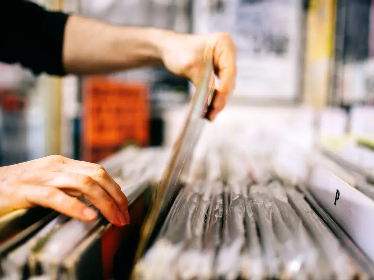 Close-up of a person’s hands flipping through a row of vinyl records in protective sleeves at a record store. The background is blurred, focusing on the action and the records.