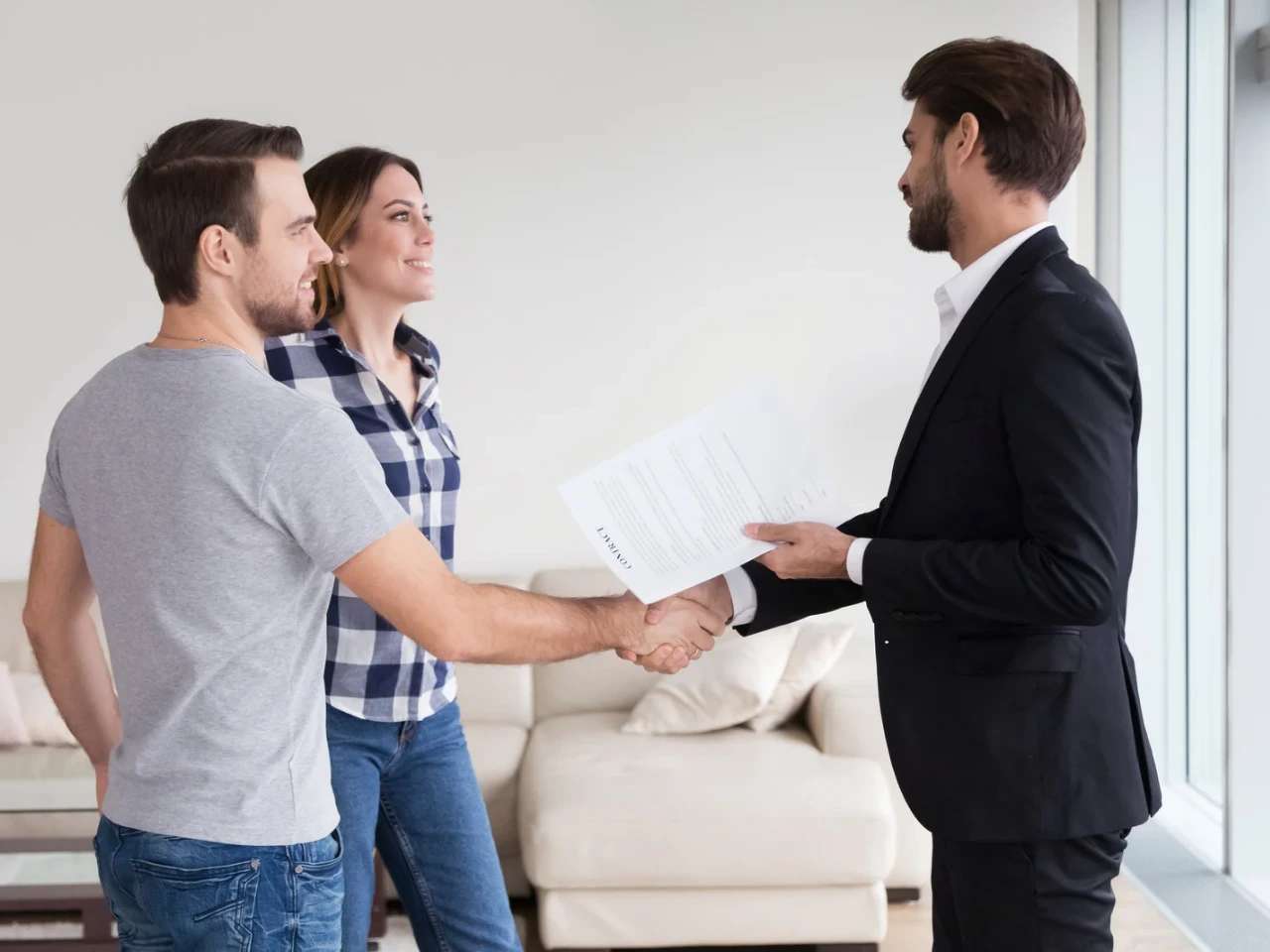 A man in a suit shakes hands with a casually dressed man while a smiling woman stands beside them in a modern living room, suggesting a successful agreement or business deal.