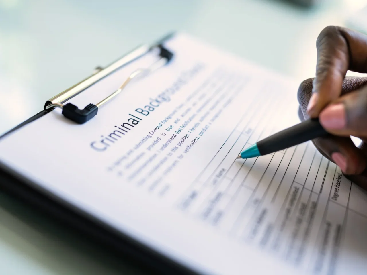 A close-up of a hand holding a pen, filling out a criminal background check form on a clipboard. The text on the form is slightly blurred but the heading is clearly visible.