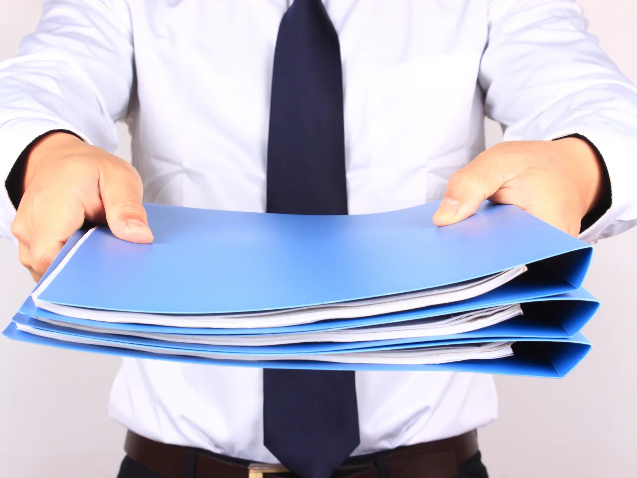 A person in a white shirt and dark tie holds out a stack of blue folders filled with papers, extending them towards the viewer.