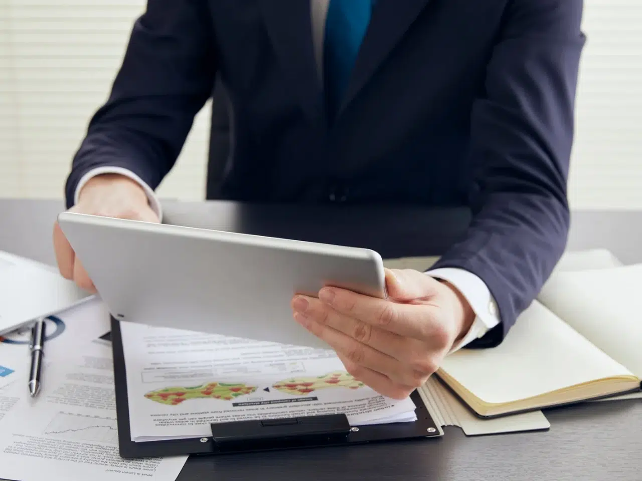 A person in a suit sits at a desk holding a tablet. There are documents with charts and a notebook on the desk. Only the persons torso and hands are visible.