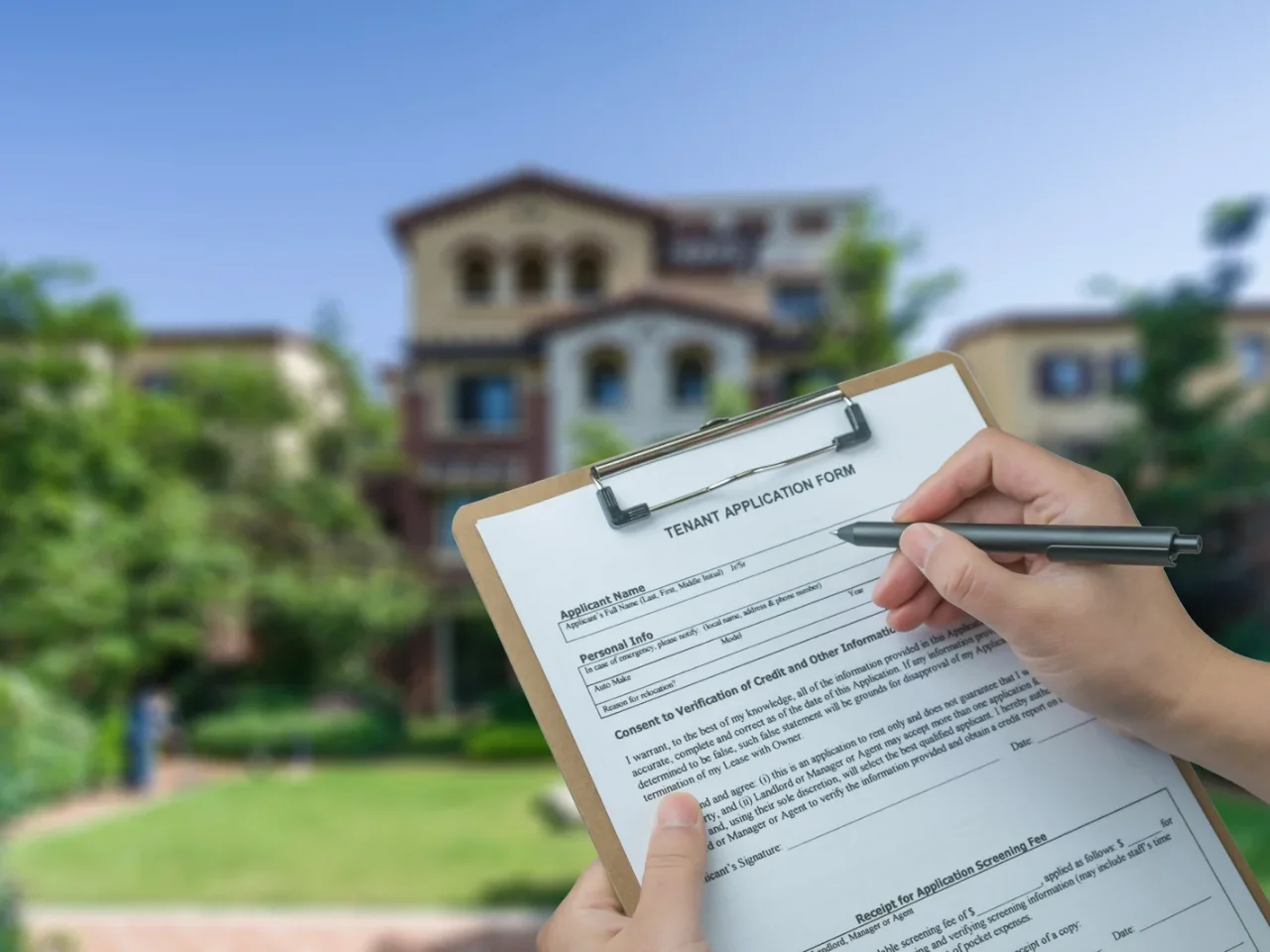 A person fills out a tenant application form on a clipboard, with a large house and green lawn blurred in the background.