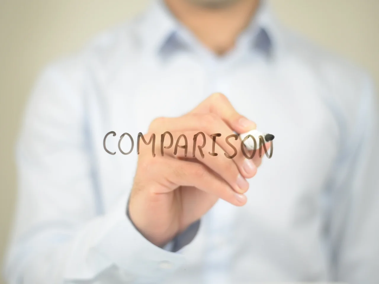 A person in a light blue shirt writes the word COMPARISON on clear glass with a black marker, with the focus on the hand and text.
