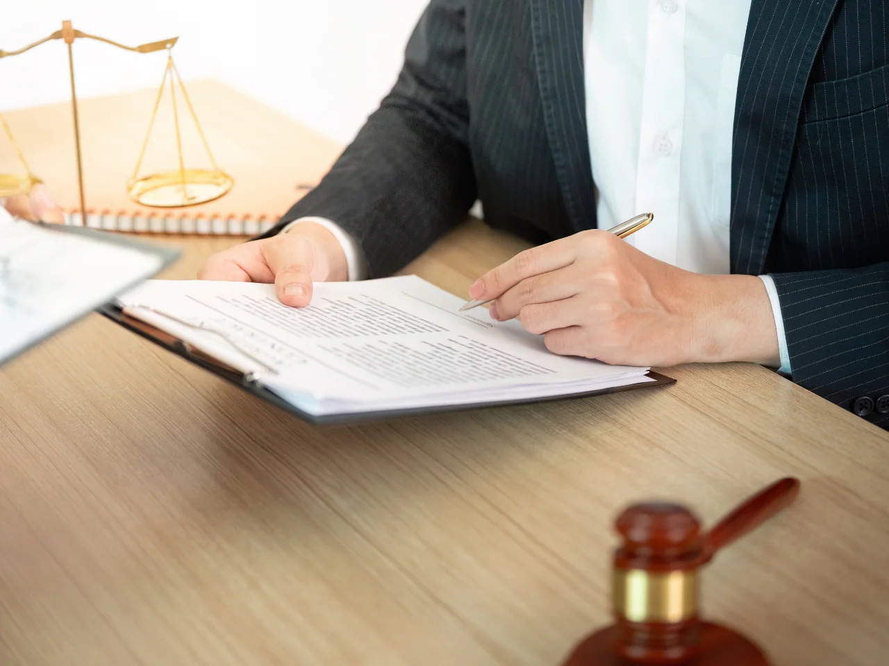 A person in a suit reviews and writes on documents at a desk with a pen, next to a gavel and a set of golden scales, suggesting a legal or courtroom setting.