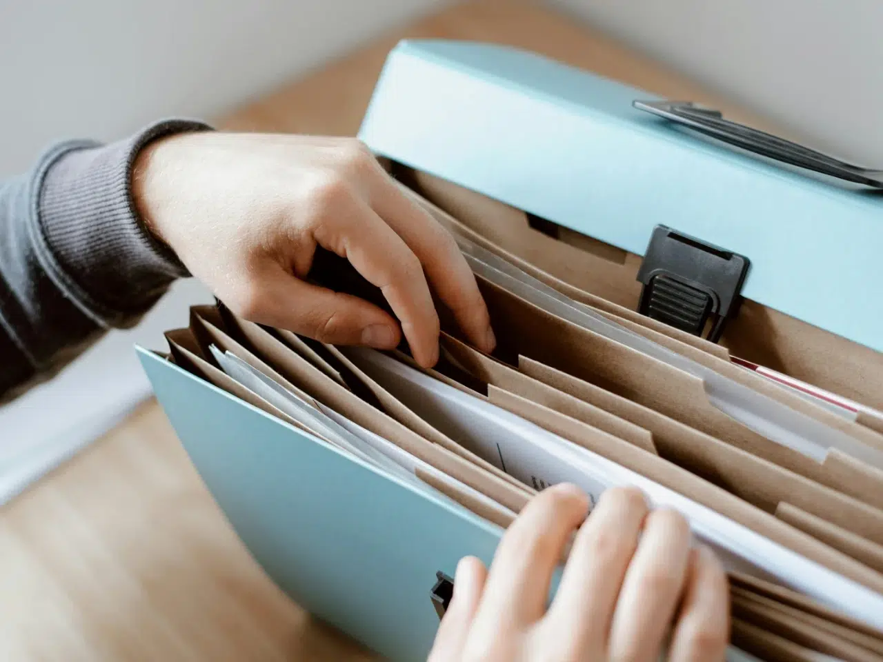 A person’s hands sorting through paperwork organized in file folders inside a light blue accordion-style folder on a wooden surface.