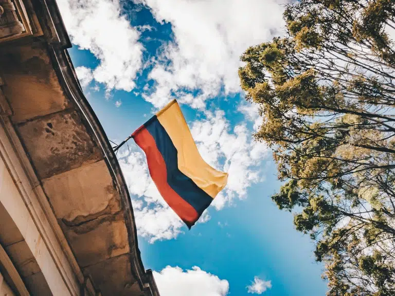 A Colombian flag waves on a pole attached to a building, with blue sky, white clouds, and tall green trees in the background.