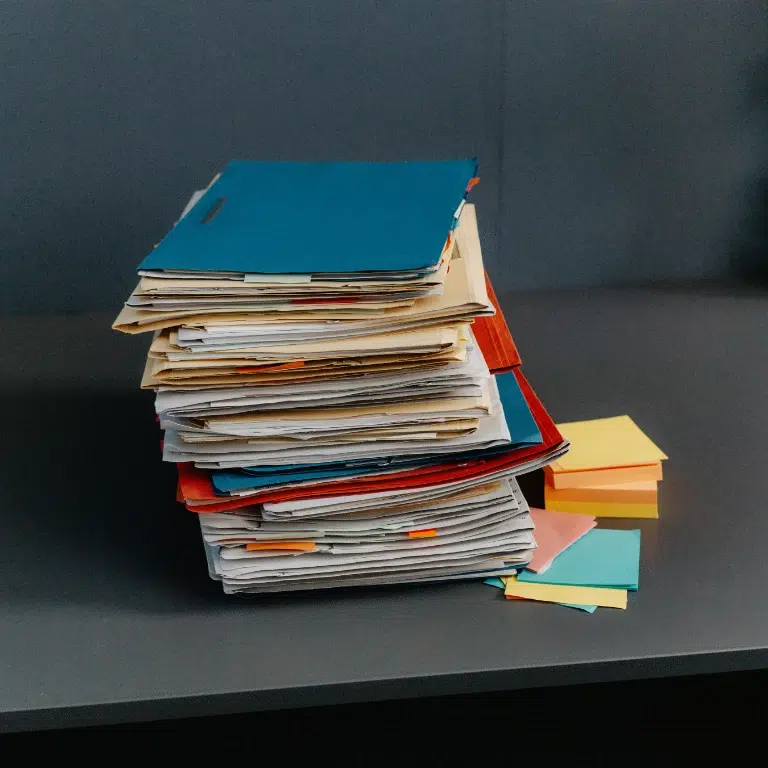 A tall stack of folders and papers sits on a gray desk, with several colorful sticky notes scattered nearby. The folders are in various colors, including blue and red, and appear to be well-used.