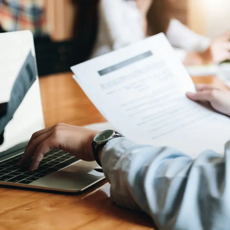 A person wearing a blue shirt and watch is holding printed documents in one hand and using a laptop with the other hand on a wooden desk, with blurred people in the background.