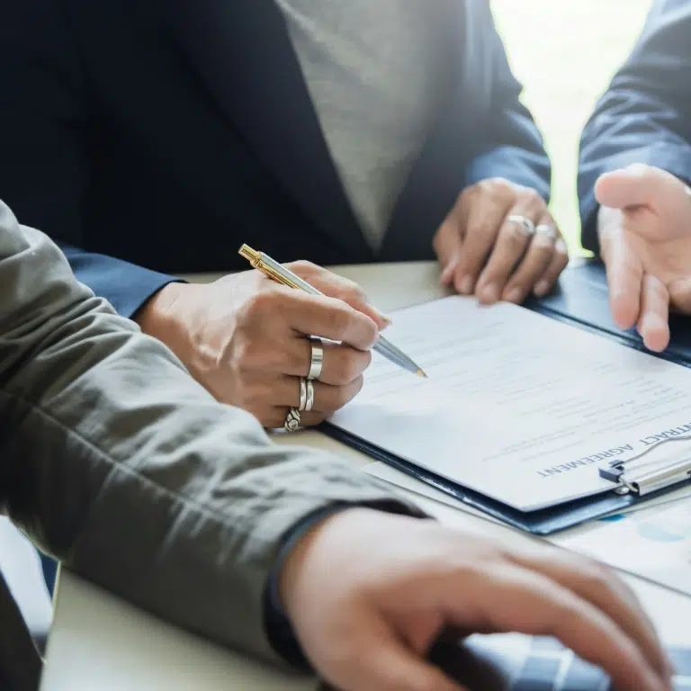Three people sit at a table. One is holding a pen and about to sign a document on a clipboard, while another points to the paper and a third types on a laptop. The scene suggests a business meeting or agreement.