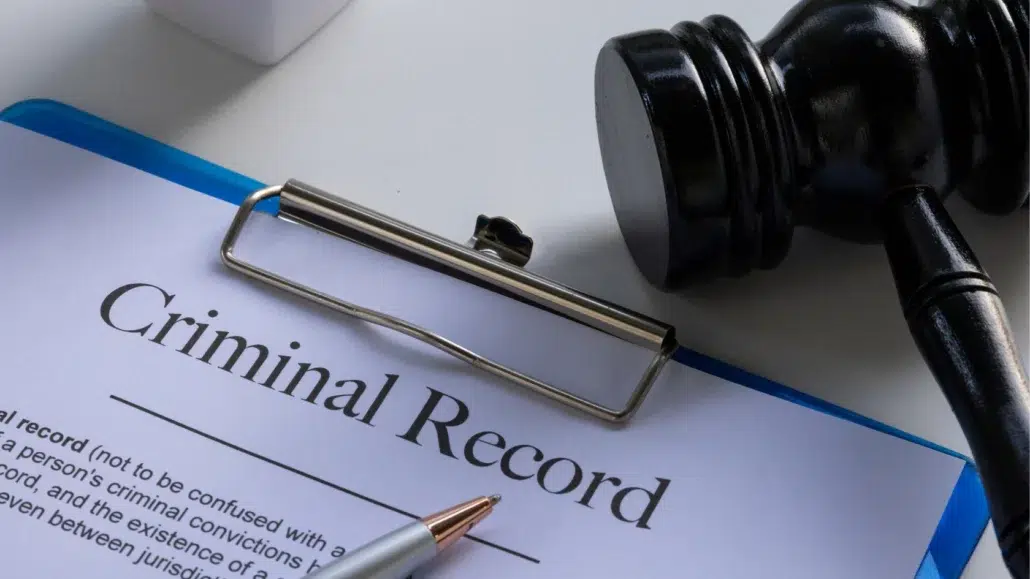 A clipboard holding a document labeled Criminal Record next to a pen and a judges gavel on a white desk.