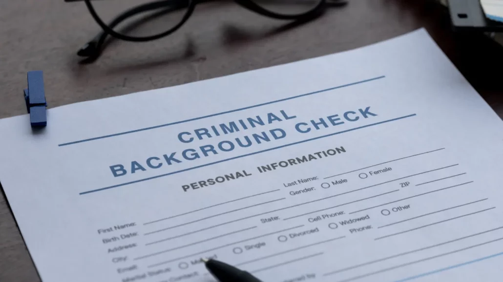 A close-up of a criminal background check form on a table, with sections for personal information. A pen, eyeglasses, and a blue paper clip are nearby. The form is partially filled out.