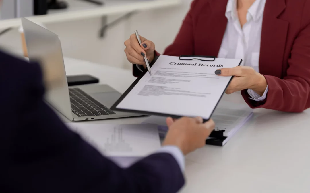 Two people in business attire sit at a desk; one hands a clipboard labeled Criminal Records to the other. A laptop and documents are visible on the desk in a professional office setting.