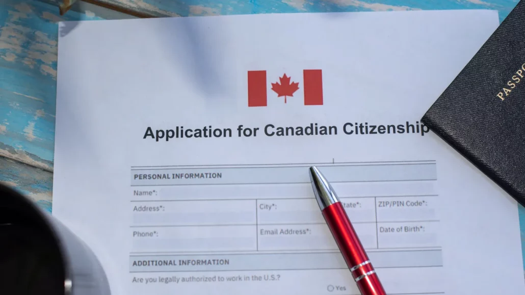 A Canadian citizenship application form with a red pen and a passport on a blue wooden surface. The top of the form features the Canadian flag.