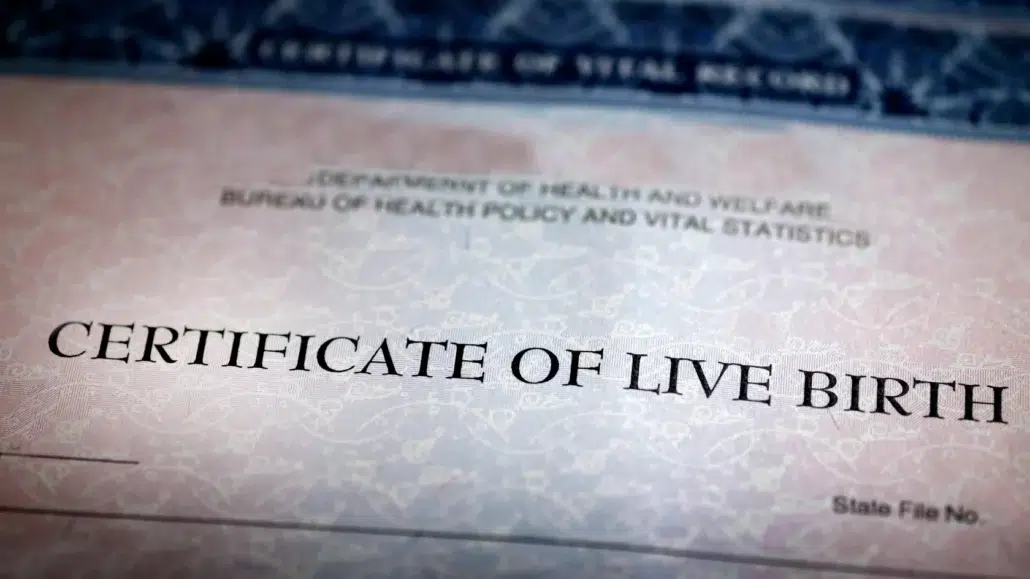 A close-up of a certificate of live birth, showing the heading in large, bold letters. The background features a decorative security pattern and partial text from a government health department.