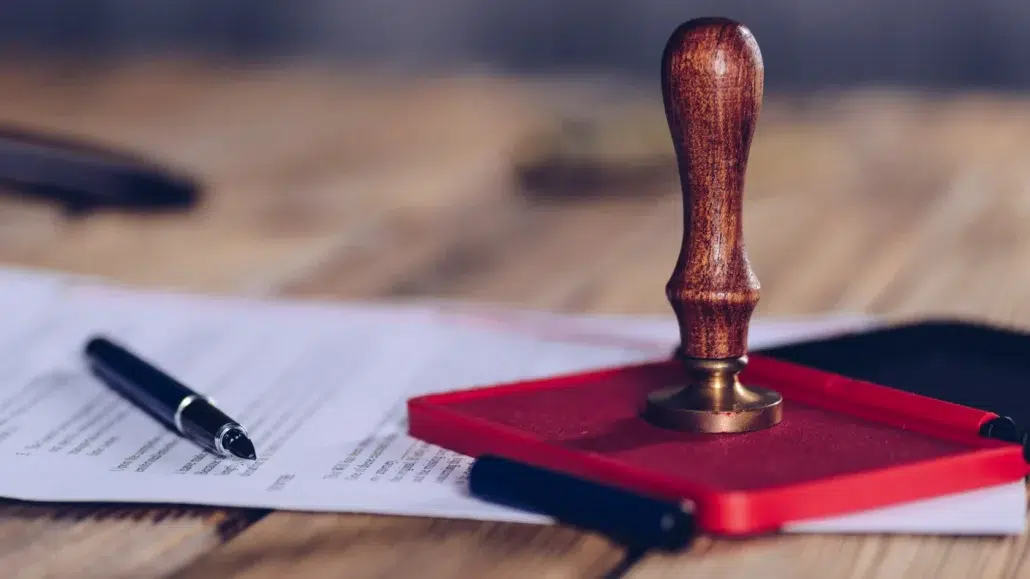 A wooden stamp on a red ink pad sits on a desk next to a few sheets of paper and two pens, suggesting the signing or stamping of official documents.