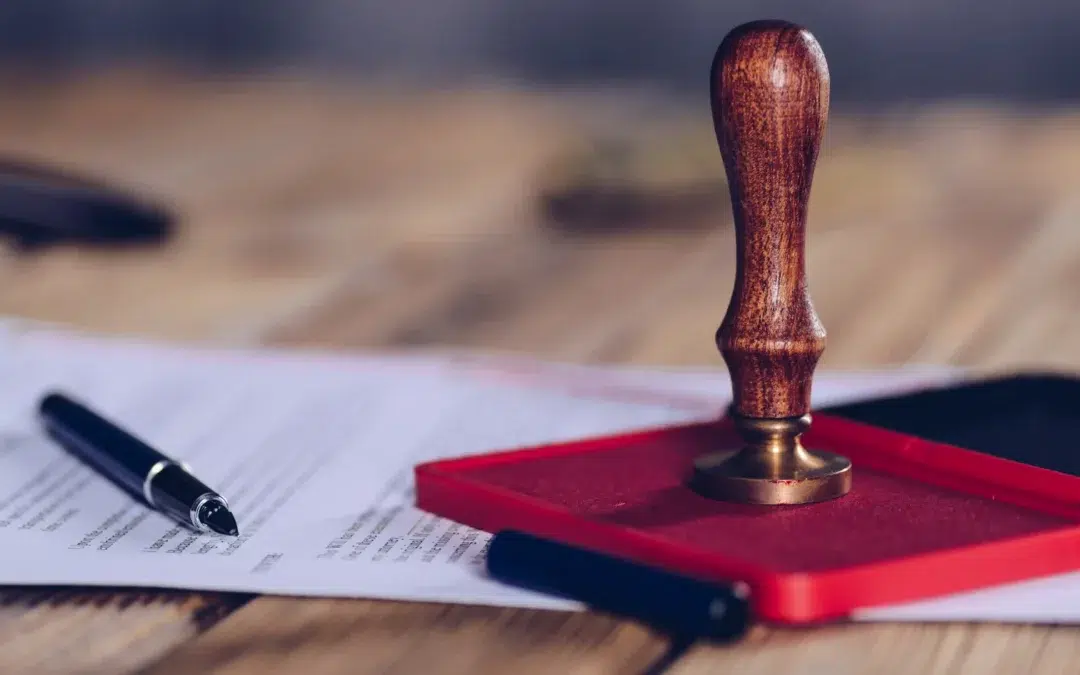 A wooden stamp on a red ink pad sits on a desk next to a few sheets of paper and two pens, suggesting the signing or stamping of official documents.