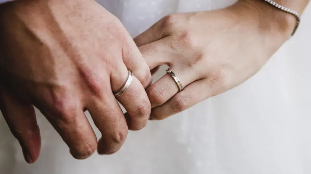 Close-up of two hands with wedding rings touching gently, likely symbolizing a newlywed couple. The background is soft and blurred, highlighting the hands and rings.