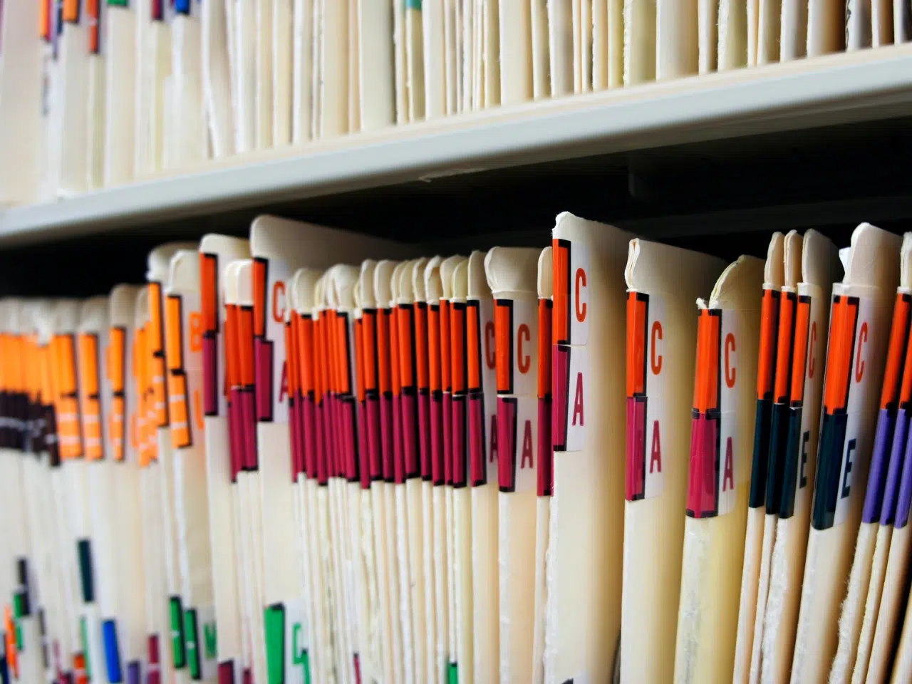 Rows of organized file folders stand upright on shelves, each labeled with colored tabs and letters, indicating an alphabetical filing system, likely used for storing documents such as medical or office records.