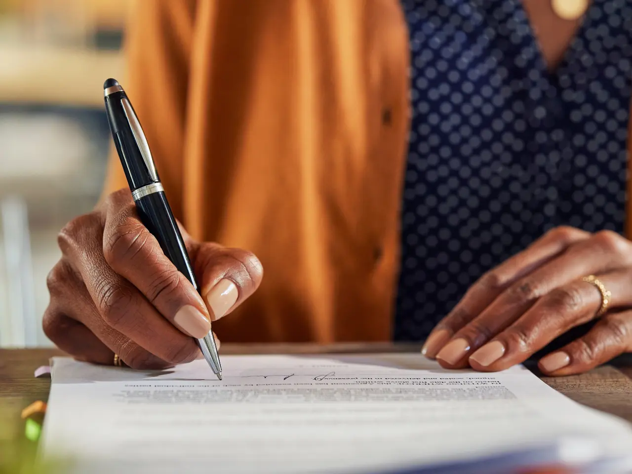 Close-up of a person’s hands signing a document with a black pen. The person is wearing a brown jacket over a blue, patterned shirt. The focus is on the hands and the pen.